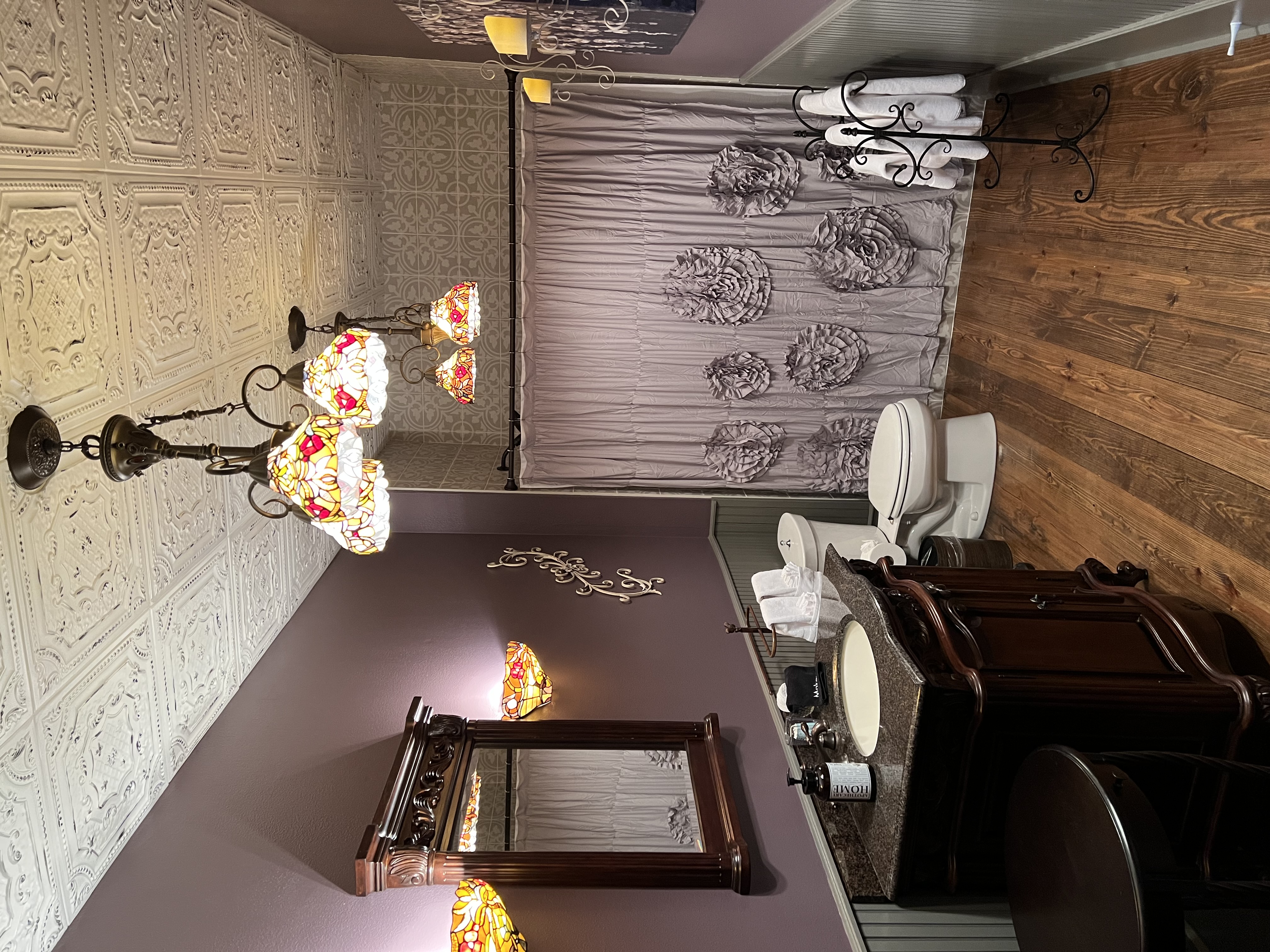 A bathroom with lavender walls, wood floors, a dark wood vanity, an ornate chandelier, floral wall sconces, and a shower with a patterned curtain.