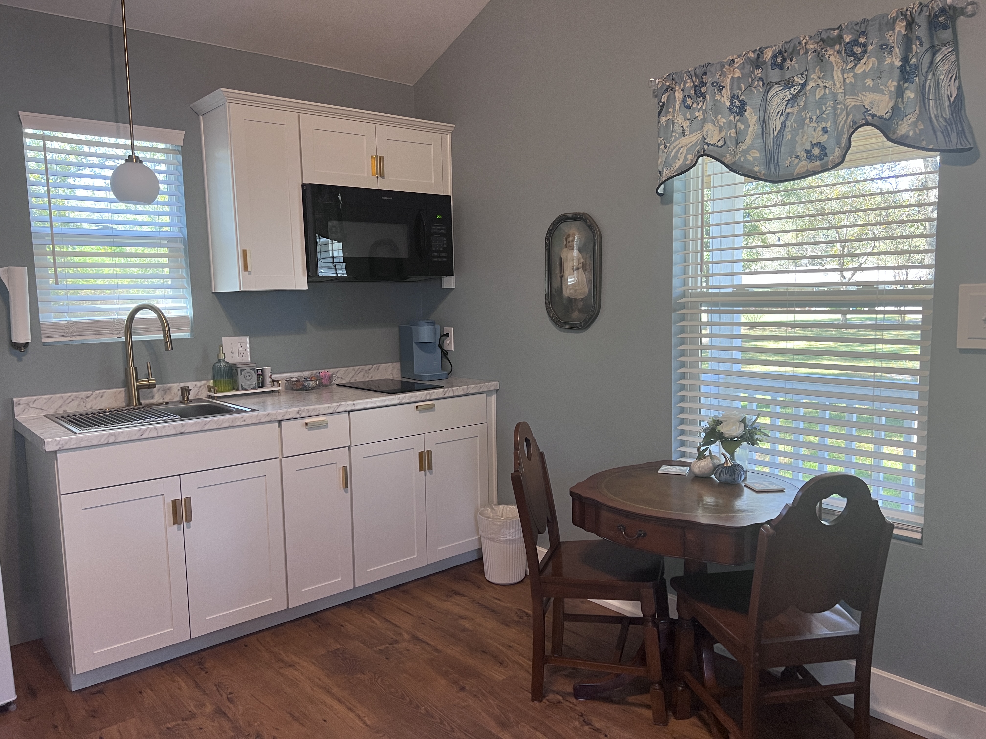 A cottage interior with light blue walls, wood floors, a sitting area with two chairs, a stacked washer/dryer, and a small kitchenette.