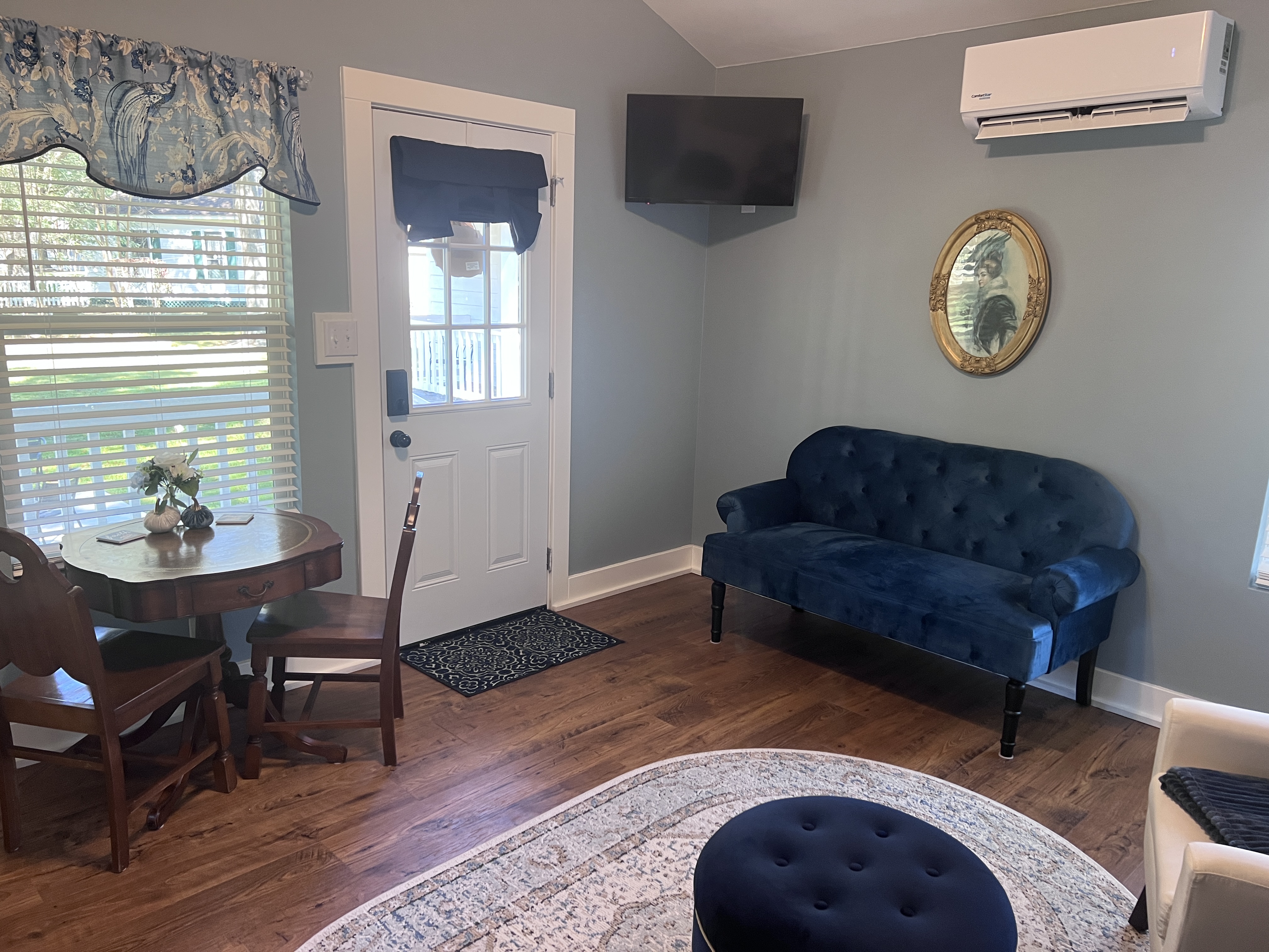 A cottage living space with light blue walls, a vaulted ceiling, a blue velvet settee, a mounted TV, a small dining table, and a door to the outside.