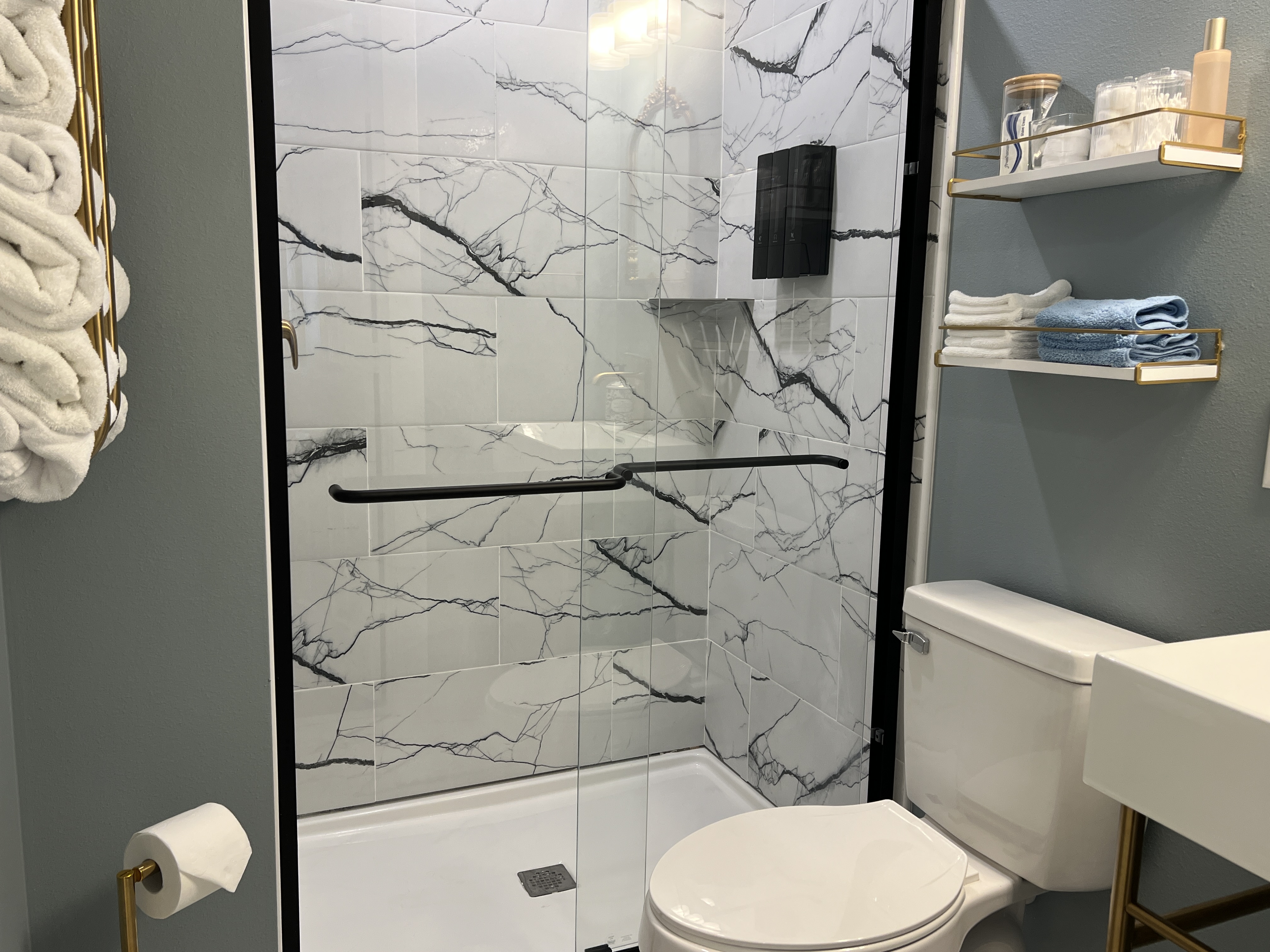 A stylish bathroom with light blue walls, a walk-in shower with marble-look tile and black accents, a modern toilet, and white shelving.
