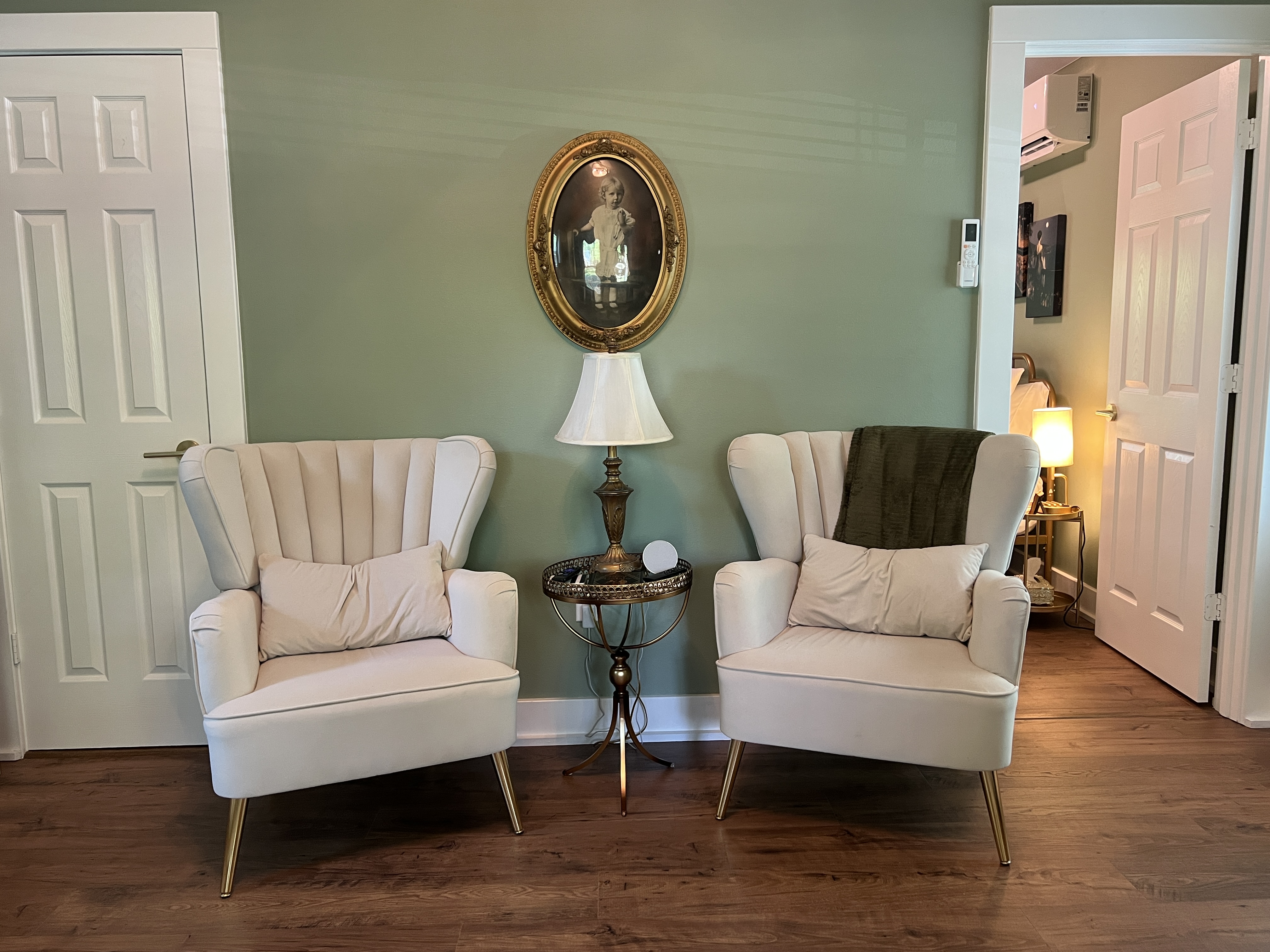 A room with sage green walls, a vaulted ceiling, two velvet wingback chairs, a stackable washer and dryer, and doorways to other rooms.