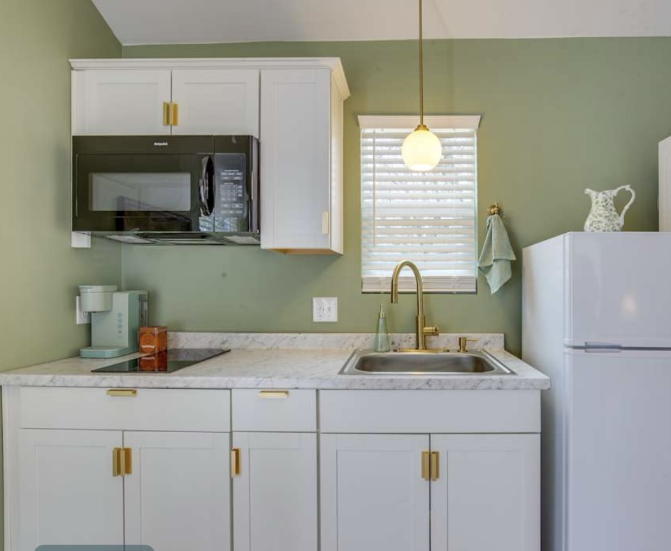 A modern kitchenette with sage green walls, white cabinets, a speckled counter, a black microwave, and gold hardware and fixtures.