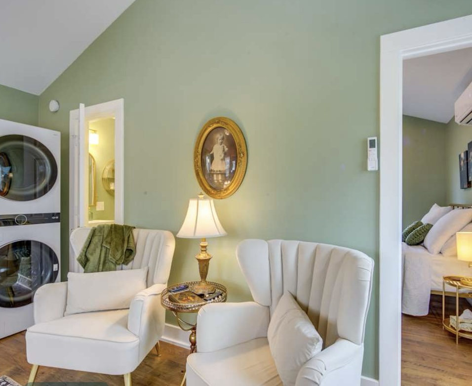 A bright sitting area with sage green walls, a vaulted ceiling, two white velvet chairs, a portrait, a stacked washer/dryer, and a view into a bedroom.