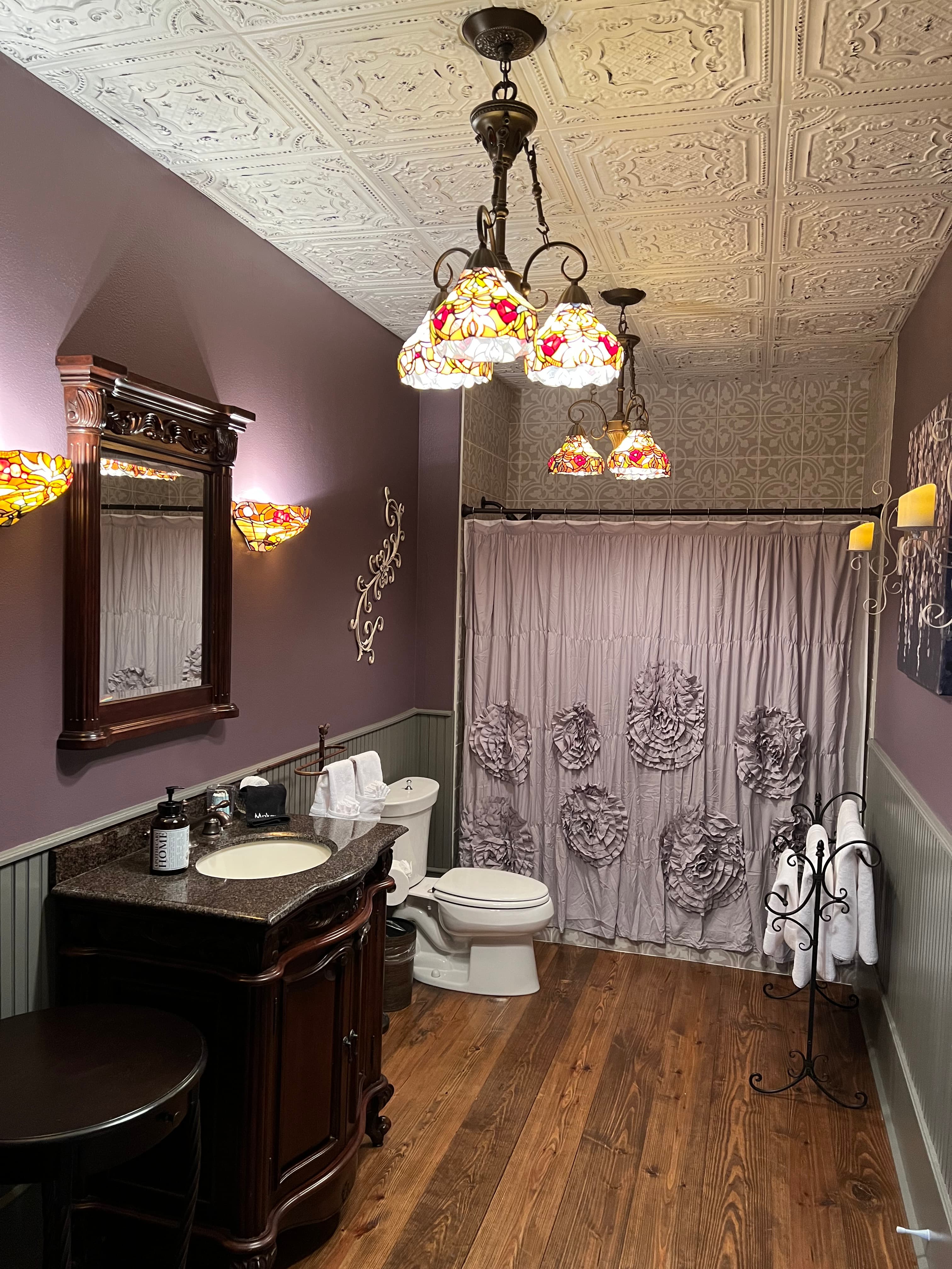 A bathroom with lavender walls, wood floors, a dark wood vanity, an ornate chandelier, floral wall sconces, and a shower with a patterned curtain.