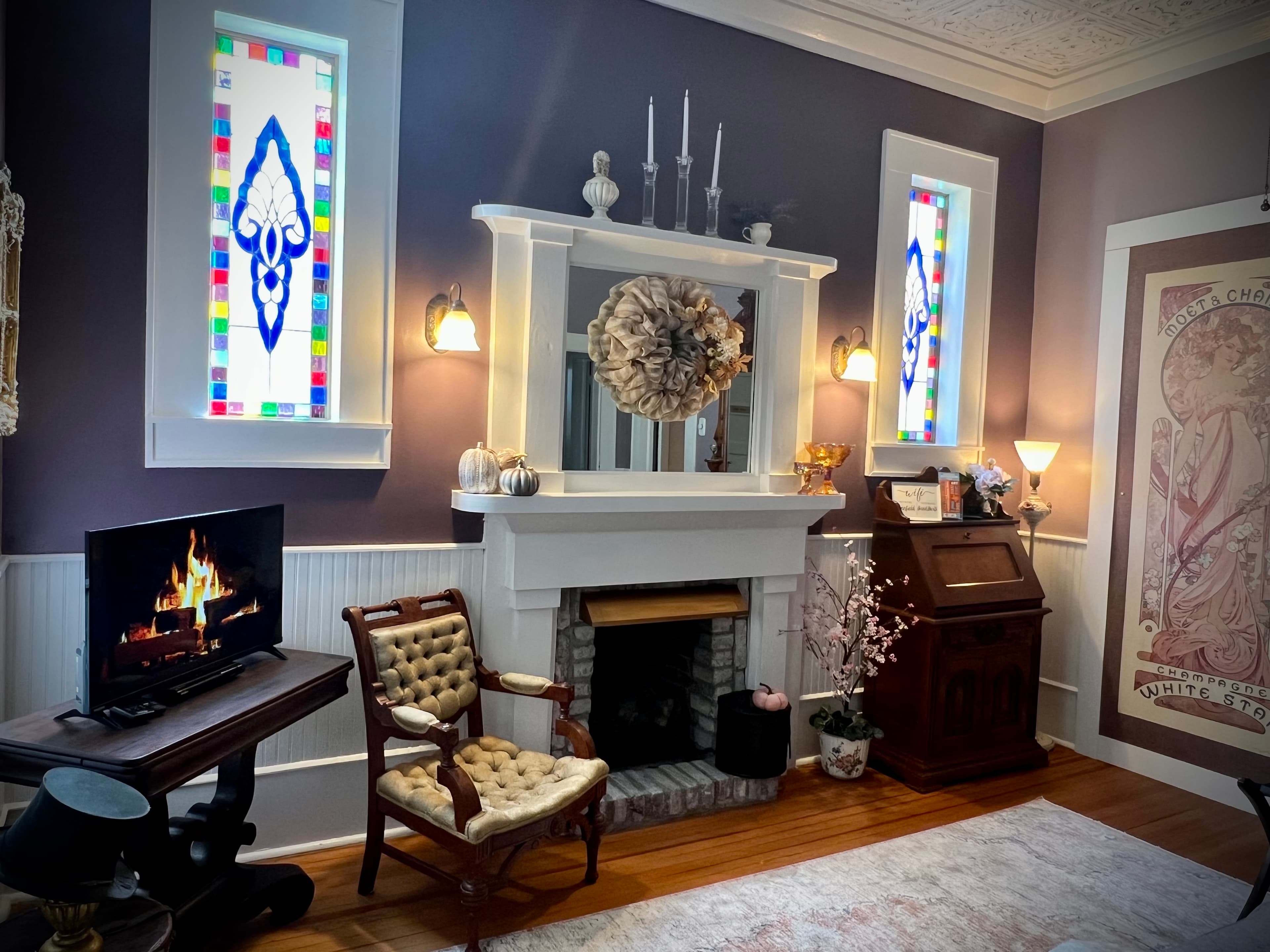 A luxurious room featuring a white fireplace mantle with a mirror, lavender walls, an electric fireplace, stained glass windows, and a patterned accent chair.