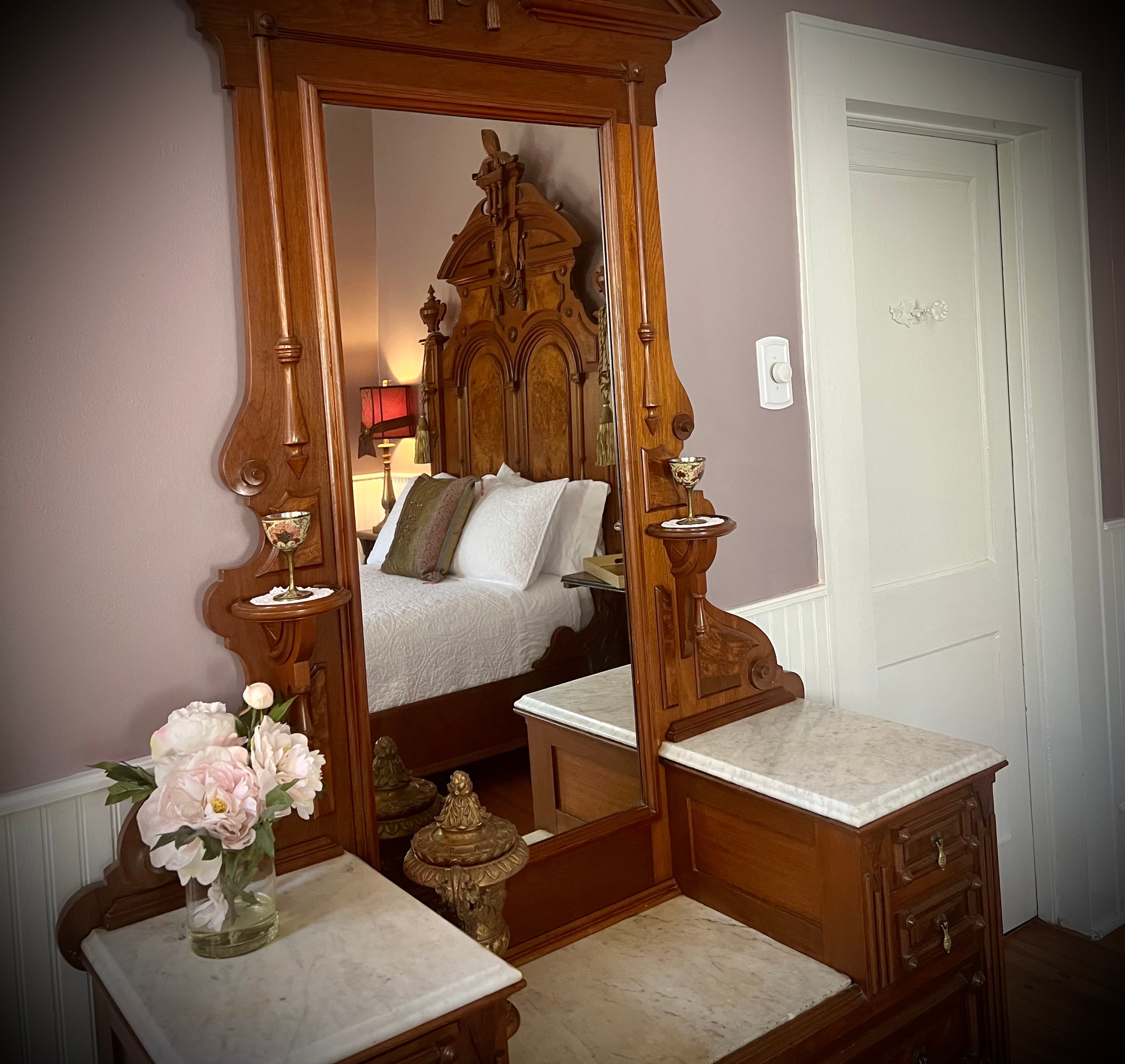 A large, ornate antique wooden marble-topped dresser with a mirror, reflecting the carved wooden headboard of a bed and a vase of pink flowers.
