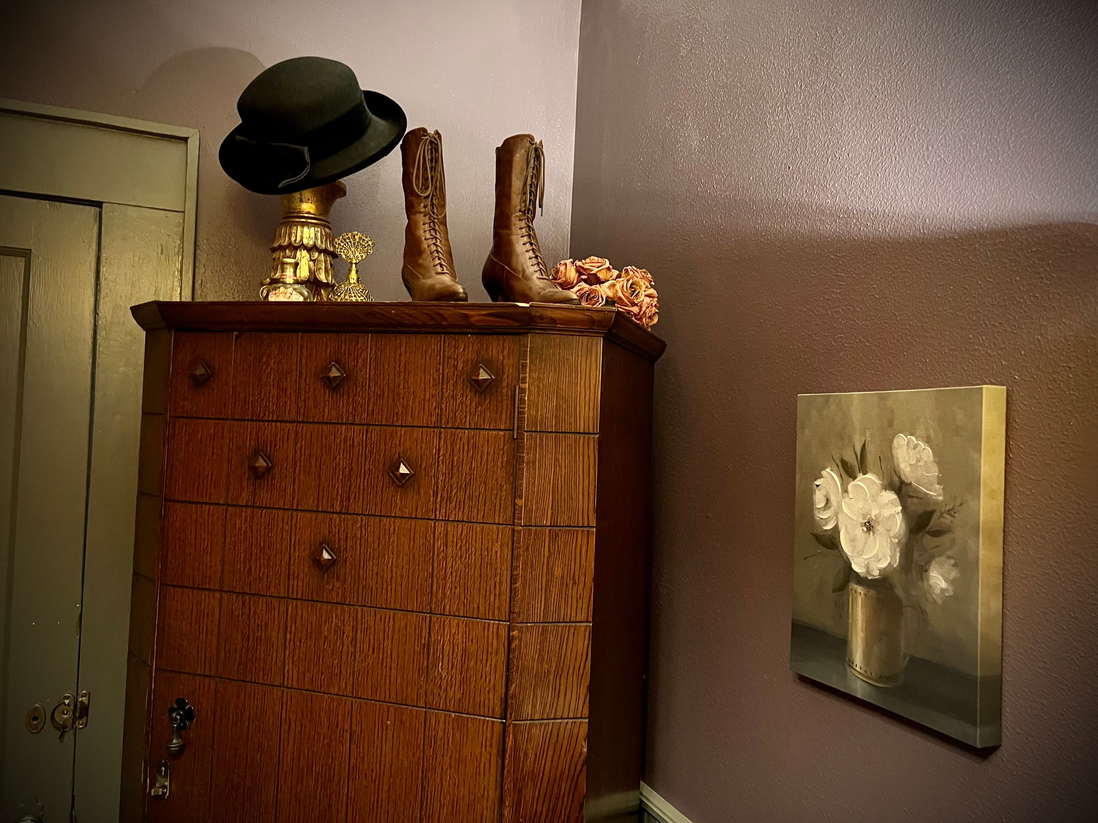 A dark wooden chest of drawers against a lavender wall, featuring Western-themed decor like cowboy boots and a fedora, with floral artwork nearby.