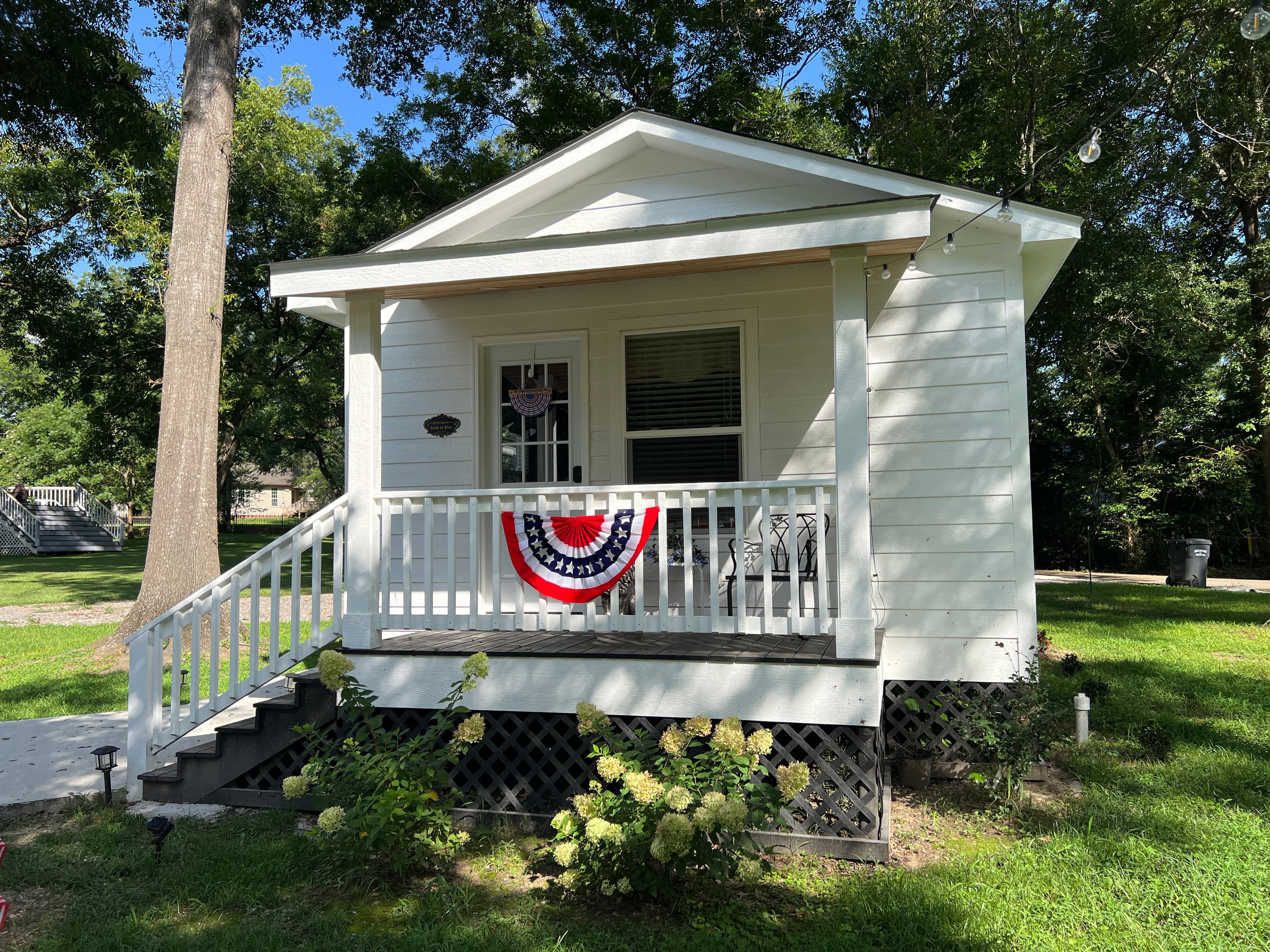 A bright white cottage with a small covered porch and steps, surrounded by green grass and trees, decorated with an American flag banner.