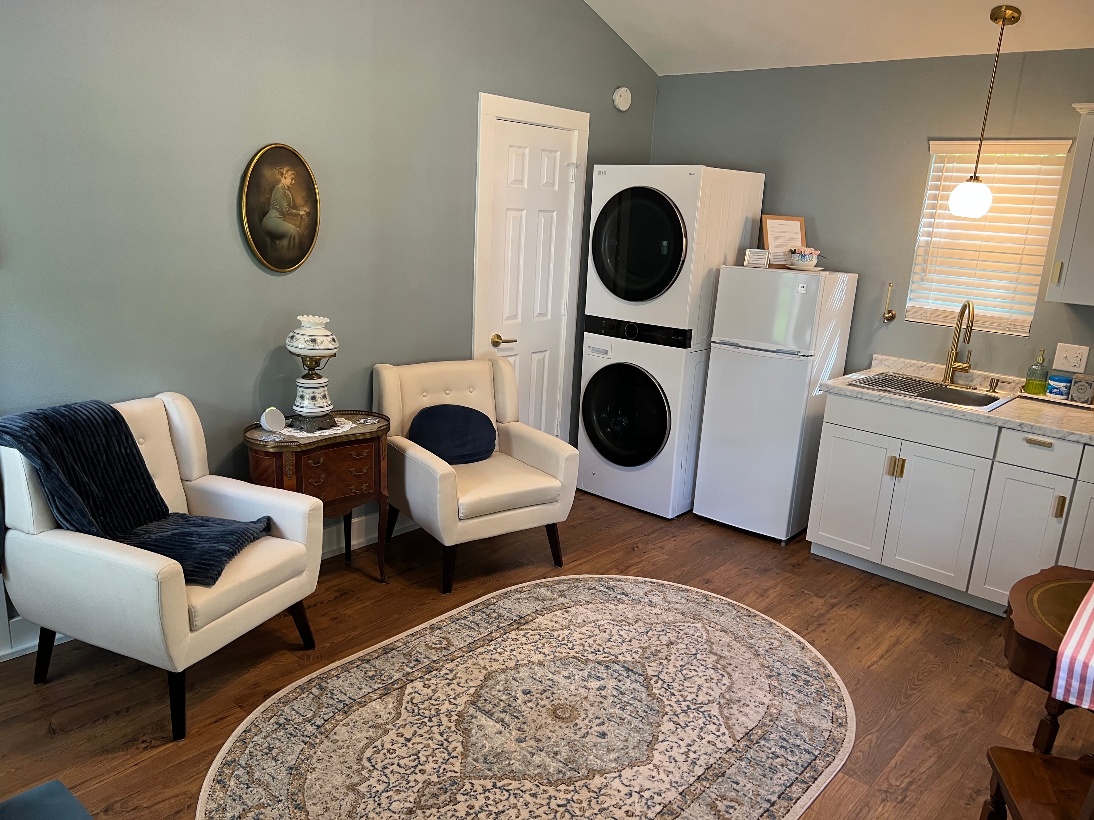 A cottage interior with light blue walls, wood floors, a sitting area with two chairs, a stacked washer/dryer, and a small kitchenette.