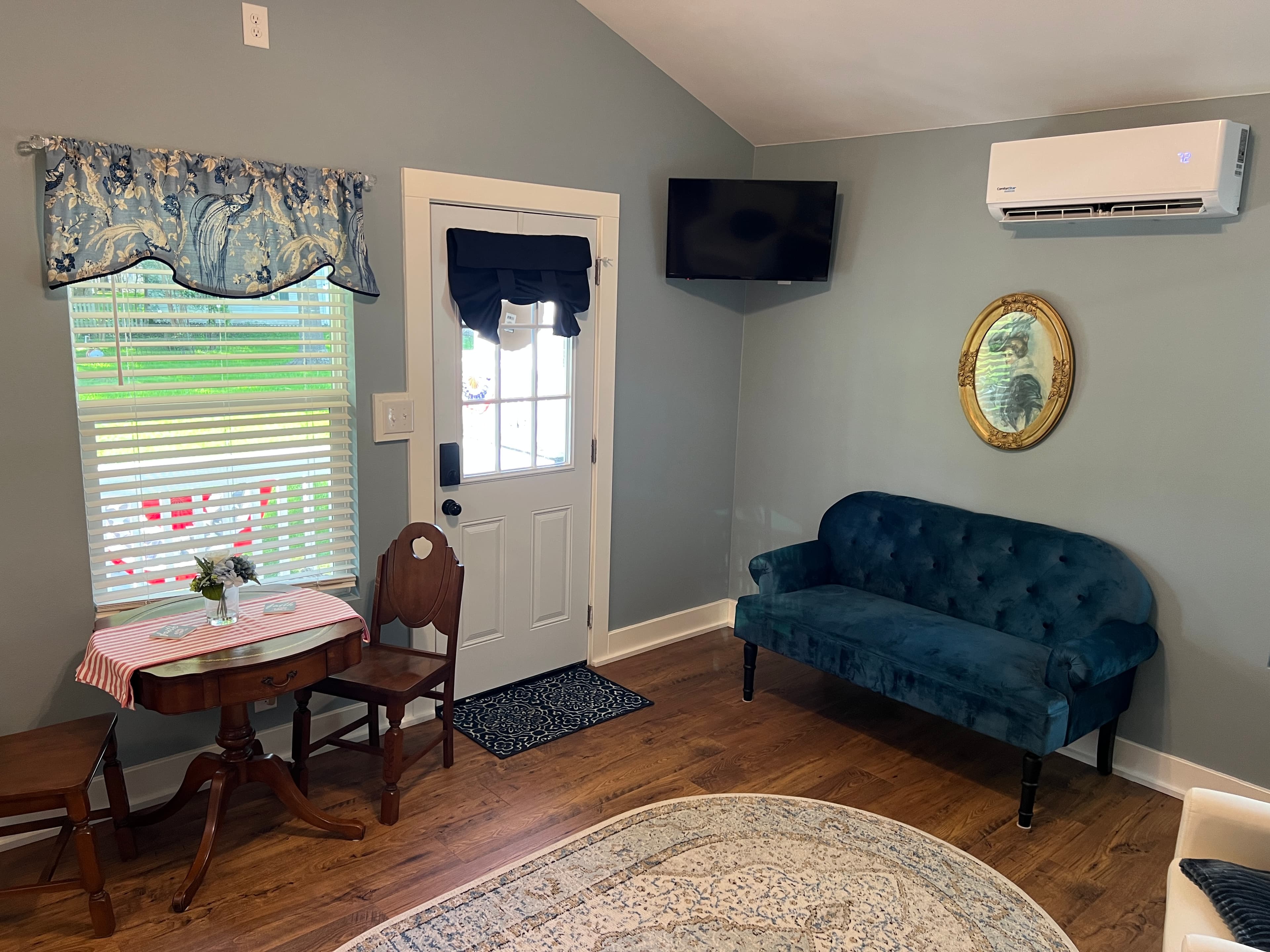A cottage living space with light blue walls, a vaulted ceiling, a blue velvet settee, a mounted TV, a small dining table, and a door to the outside.