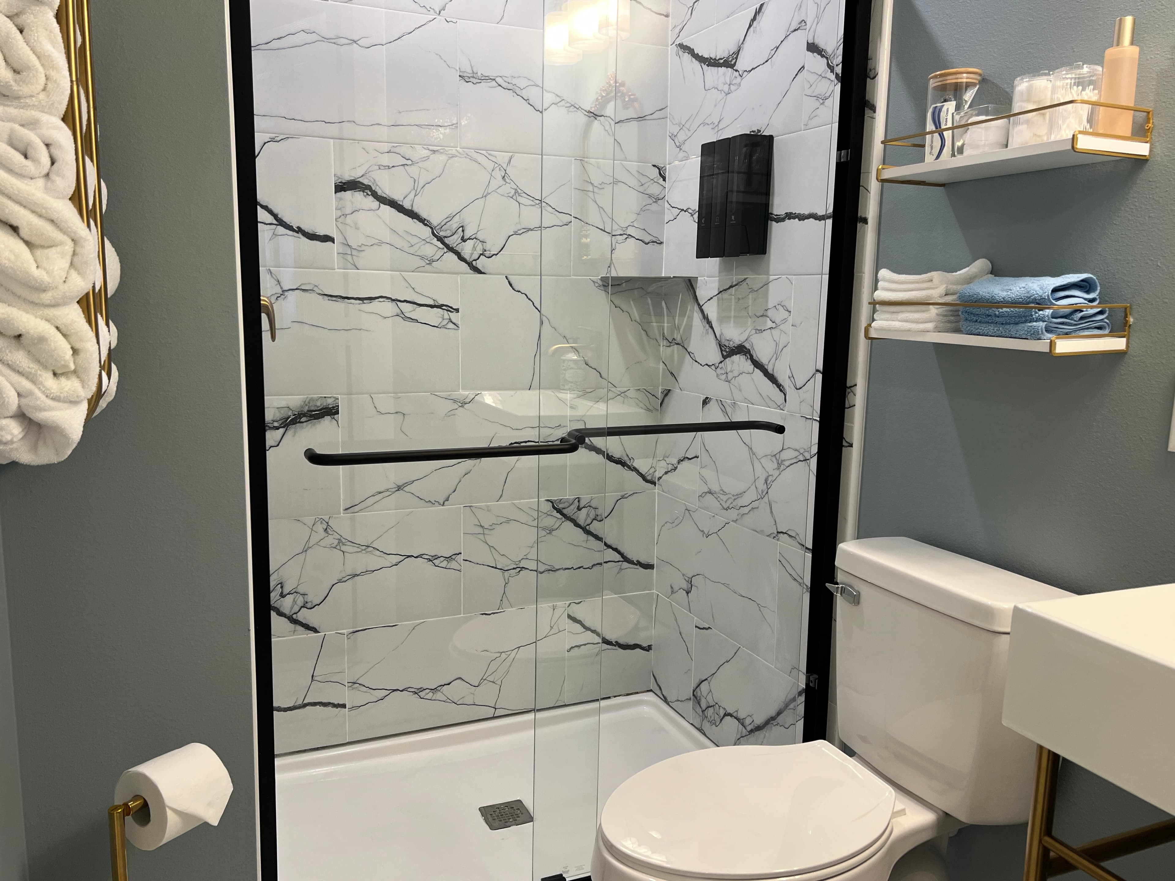 A stylish bathroom with light blue walls, a walk-in shower with marble-look tile and black accents, a modern toilet, and white shelving.