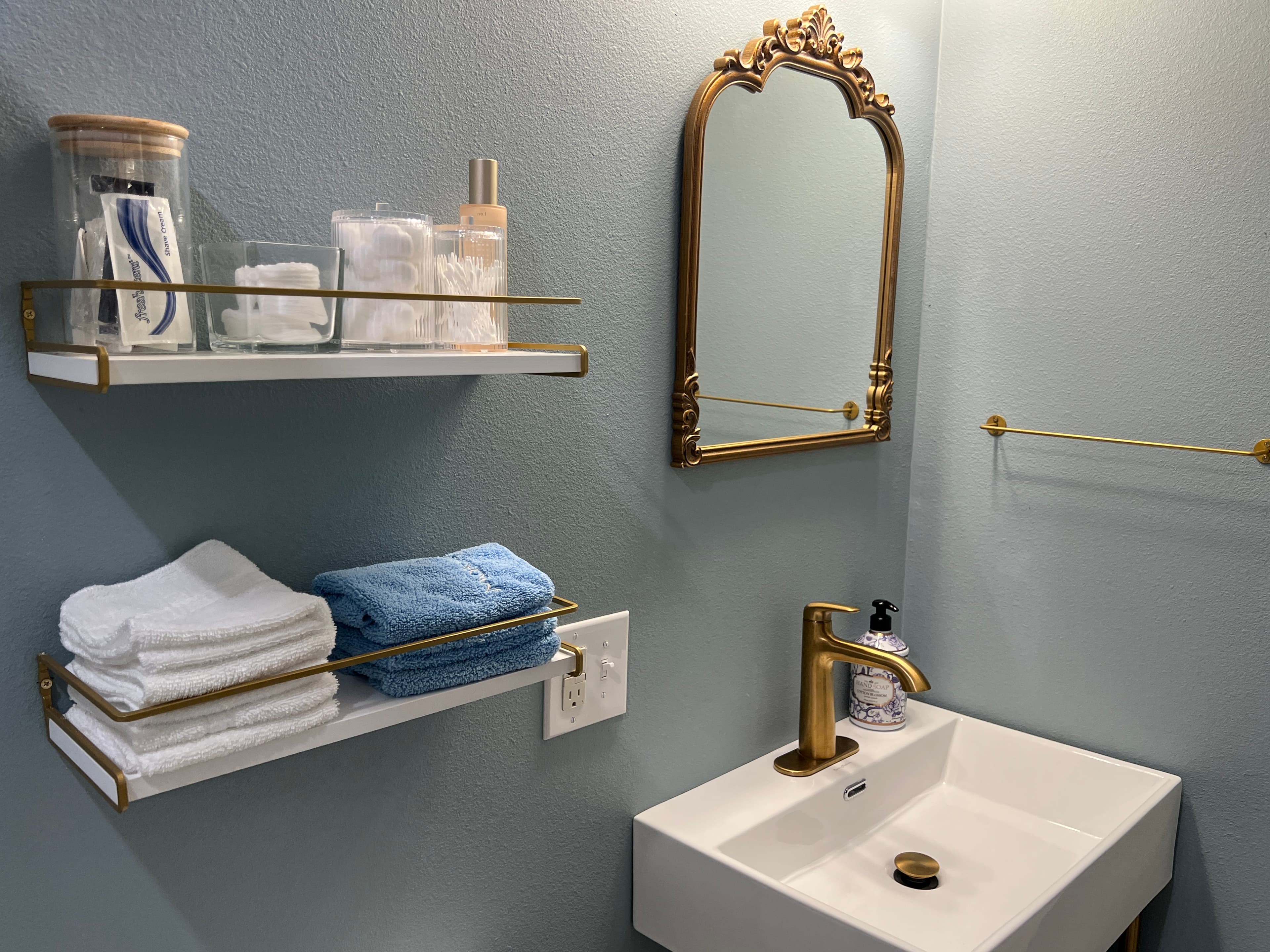 A charming vanity area with light blue walls, a white floating sink, gold accents, an ornate gold mirror, and two stacked white storage shelves.