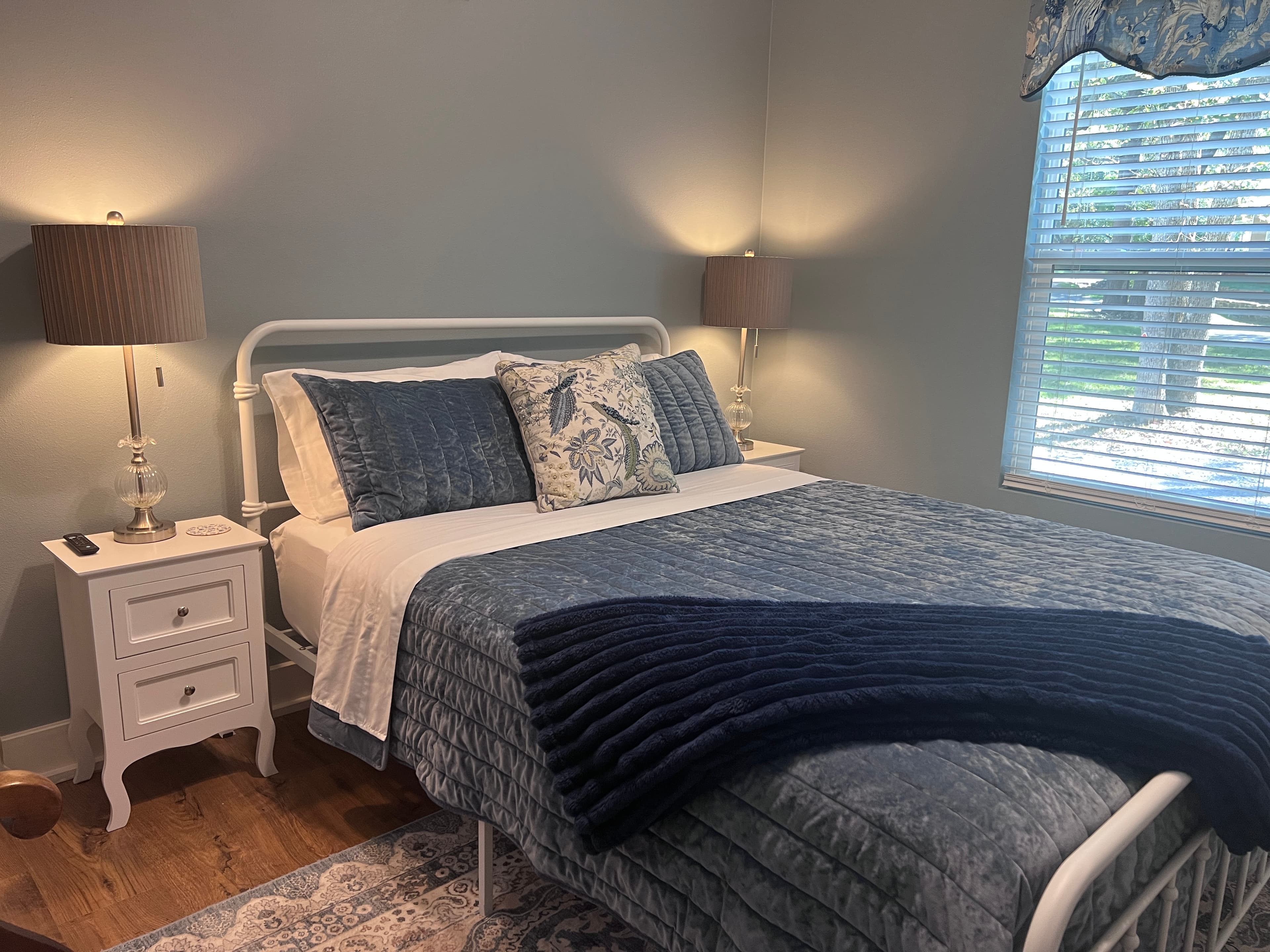 A bright, cozy bedroom with light blue walls, a white metal bed frame, matching nightstands, a patterned rug, and navy blue and white bedding.