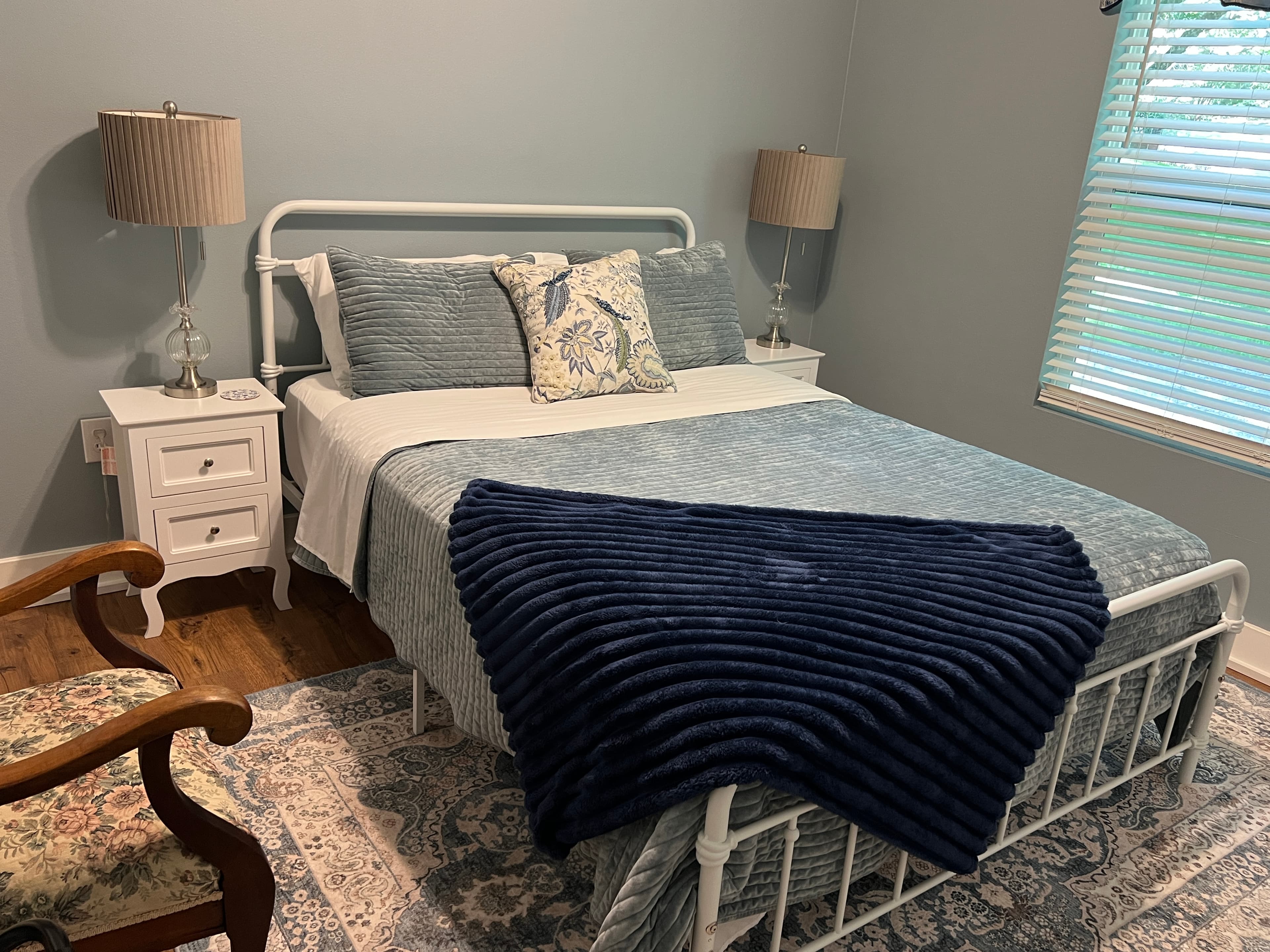 A bright, cozy bedroom with light blue walls, a white metal bed frame, matching nightstands, a patterned rug, and navy blue and white bedding.