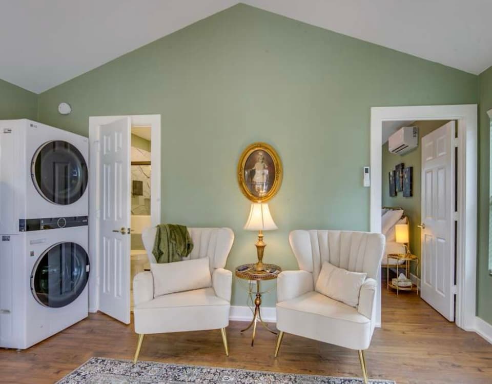 A room with sage green walls, a vaulted ceiling, two velvet wingback chairs, a stackable washer and dryer, and doorways to other rooms.