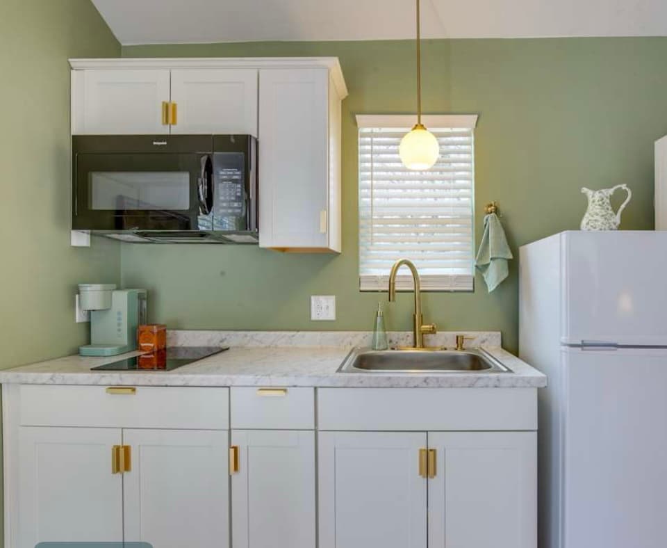 A modern kitchenette with sage green walls, white cabinets, a speckled counter, a black microwave, and gold hardware and fixtures.