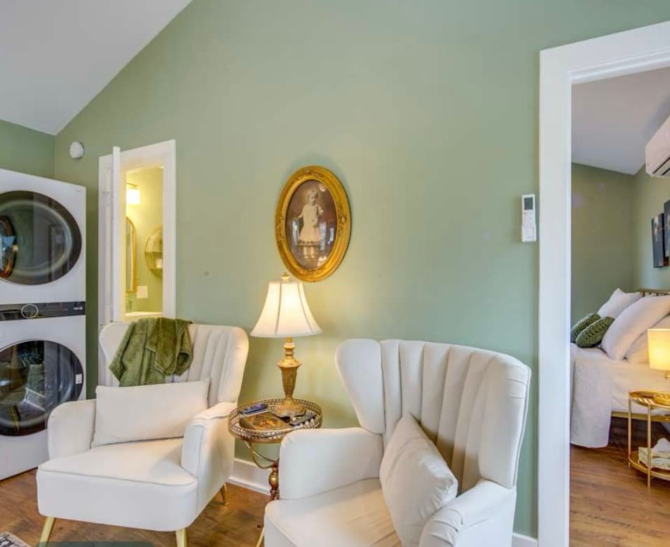 A bright sitting area with sage green walls, a vaulted ceiling, two white velvet chairs, a portrait, a stacked washer/dryer, and a view into a bedroom.