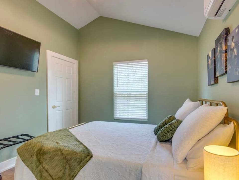 A cozy bedroom with sage green walls, a vaulted ceiling, a queen bed with white linens and green accents, a mounted TV, and a small window.