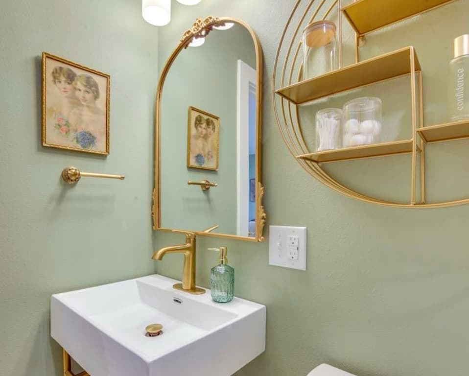 A chic bathroom with sage green walls, a white floating sink, an arched gold mirror, a matching gold faucet and towel ring, and circular shelving.