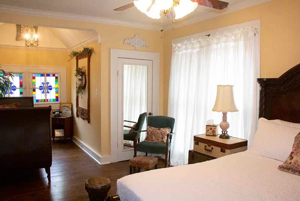 A sunny bedroom with soft yellow walls, a dark wood bed, sheer white curtains, and a seating area with stained glass windows and a private exit.