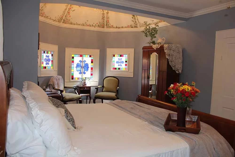 A bright, luxurious bedroom with a large bed, light blue walls, a domed ceiling, and an alcove with a seating area and stained glass windows.