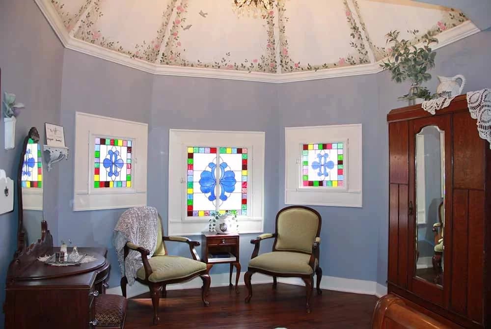A beautiful alcove with light blue walls, two vintage chairs, a small table, stained glass windows, and a hand-painted domed ceiling.