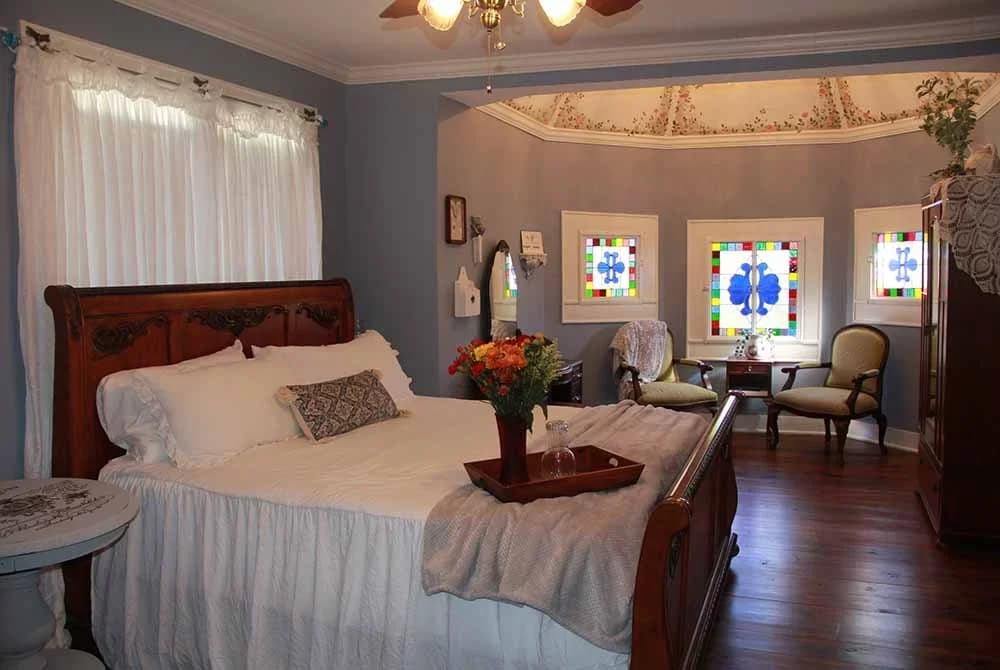 A bright, elegant bedroom with a sleigh bed, sheer curtains, and a rounded alcove featuring a seating area, stained glass windows, and a domed ceiling.
