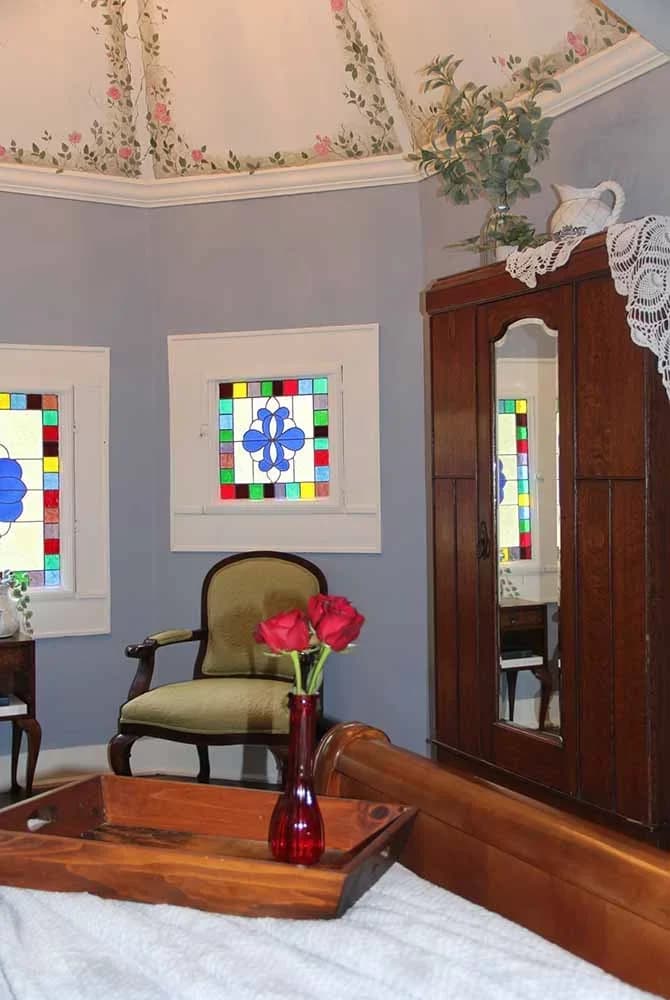 A bedroom corner with a vintage armoire and velvet armchair, set against light blue walls with stained glass windows and a hand-painted ceiling.