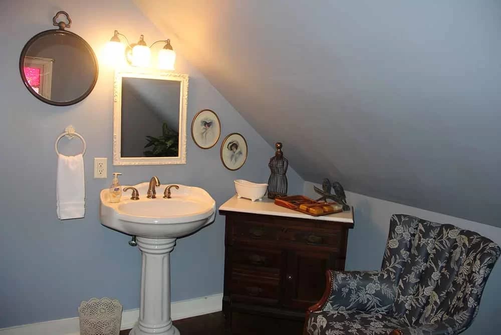 A small bathroom/vanity area with light blue walls, a pedestal sink, an accent chair, a wooden chest, and an angled ceiling from the roofline.