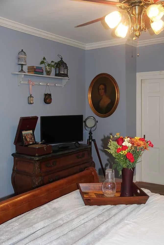A bright bedroom corner with light blue walls, a wooden chest/TV stand, a ceiling fan, a round portrait, and a tray of fresh flowers on the bed.