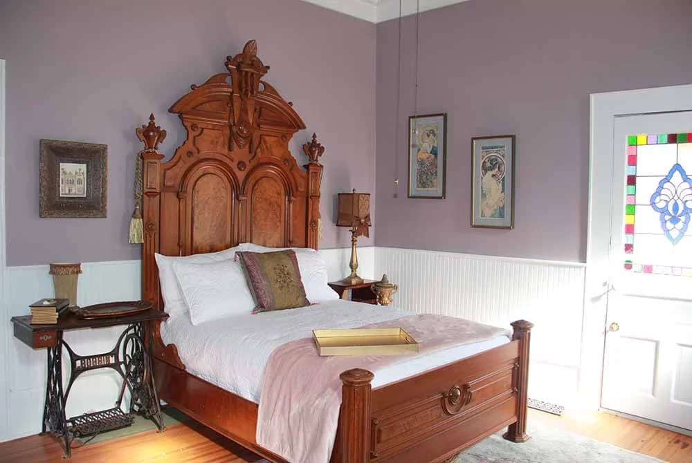 A bedroom featuring an antique wooden bed with a tall, ornate headboard, light purple walls, a vintage sewing machine table, and a door with stained glass.