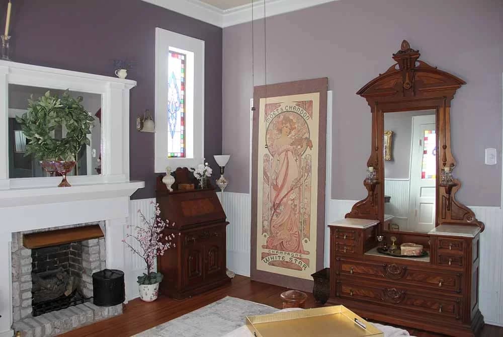 An elegantly decorated room with lavender walls, a white fireplace, an antique secretary desk, a tall wooden dresser with a mirror, and stained glass windows.