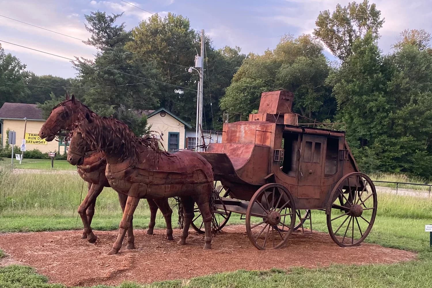 A rusted metal sculpture of two horses pulling a vintage carriage in a grassy field.