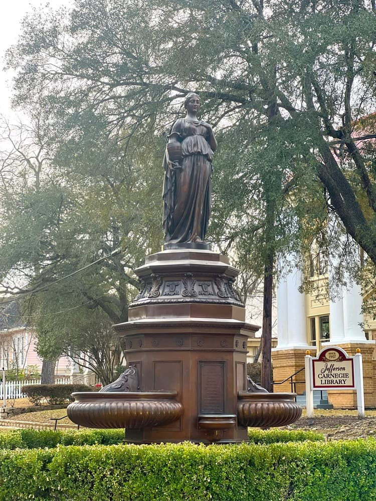 A bronze statue of a woman stands atop a fountain, surrounded by trees and near the Jefferson Carnegie Library.