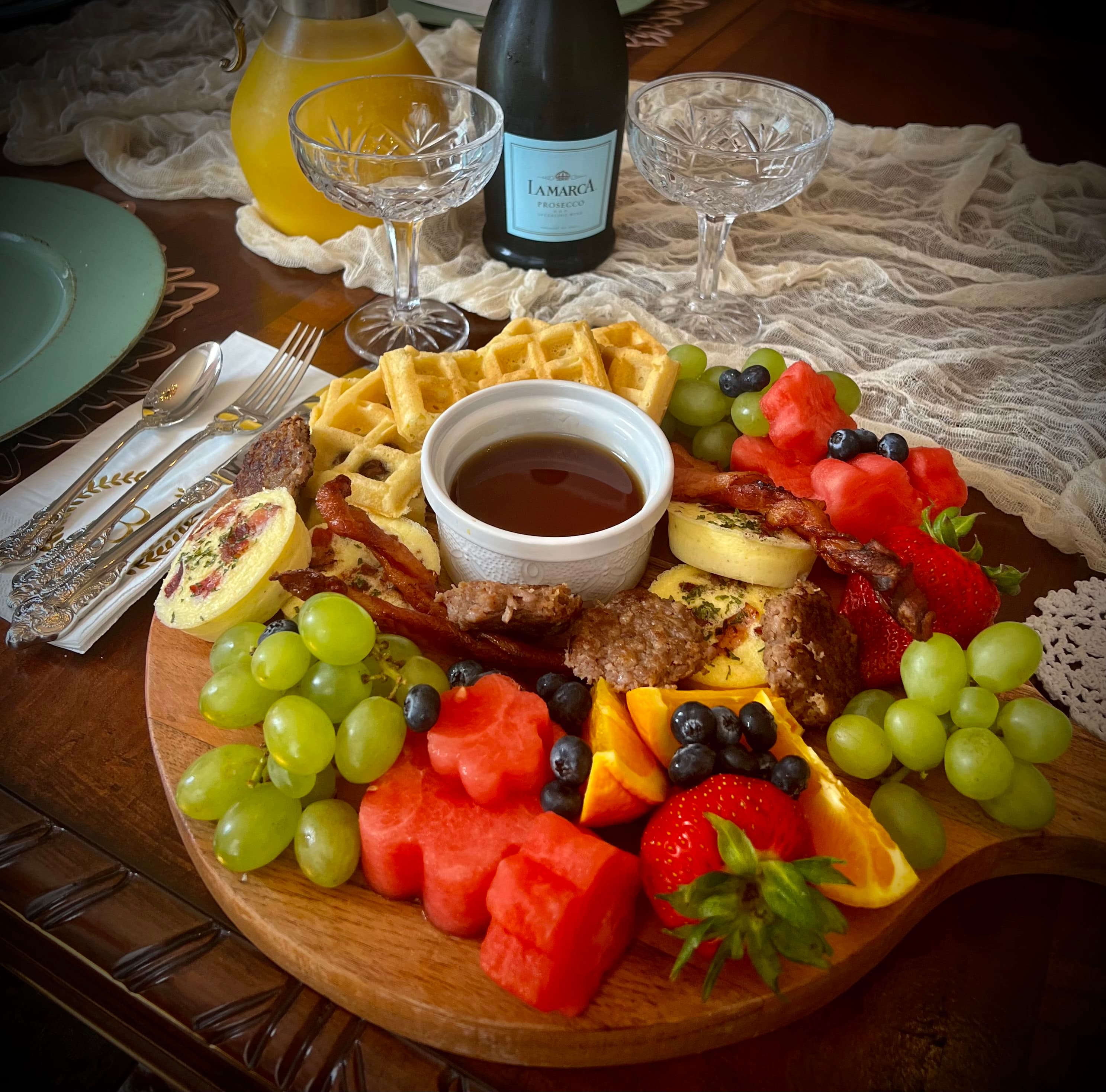 A beautifully arranged brunch platter featuring waffles, fresh fruit, bacon, and a small dish of syrup, accompanied by sparkling wine in elegant glasses.