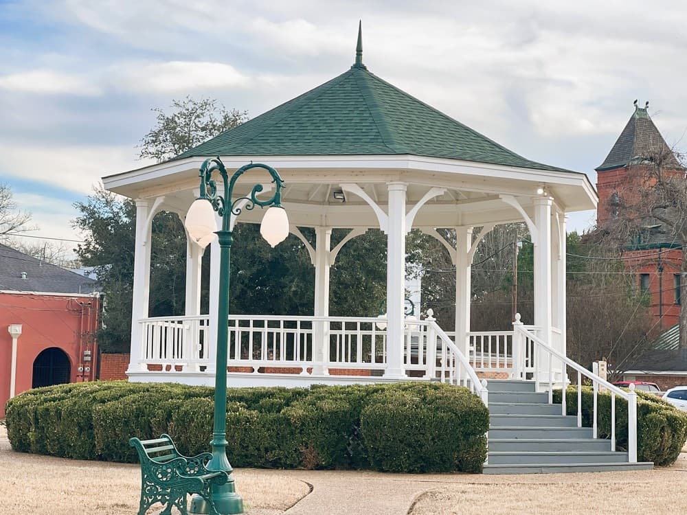 A white gazebo with a green roof surrounded by neatly trimmed bushes and a decorative street lamp.