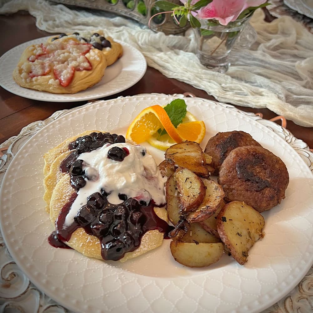 A plate of pancakes topped with blueberries and whipped cream, accompanied by seasoned potatoes, sausage, and garnished with orange slices and mint.