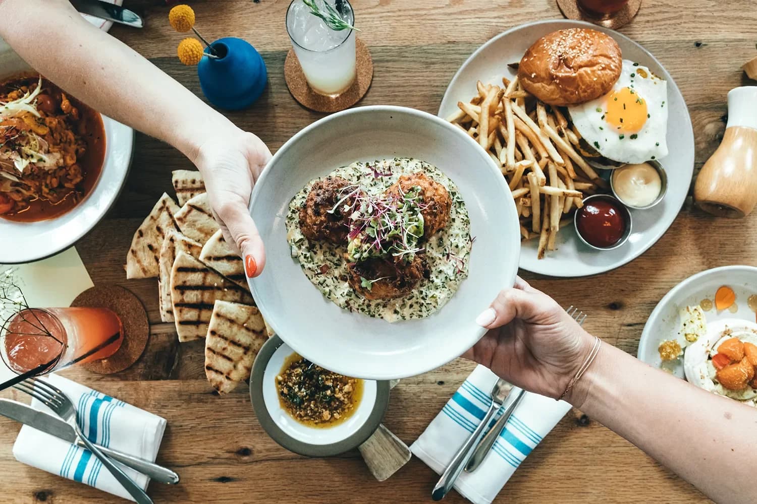 A hand reaches out to offer a plate of rice and meatballs amidst a spread of various dishes on a wooden table.