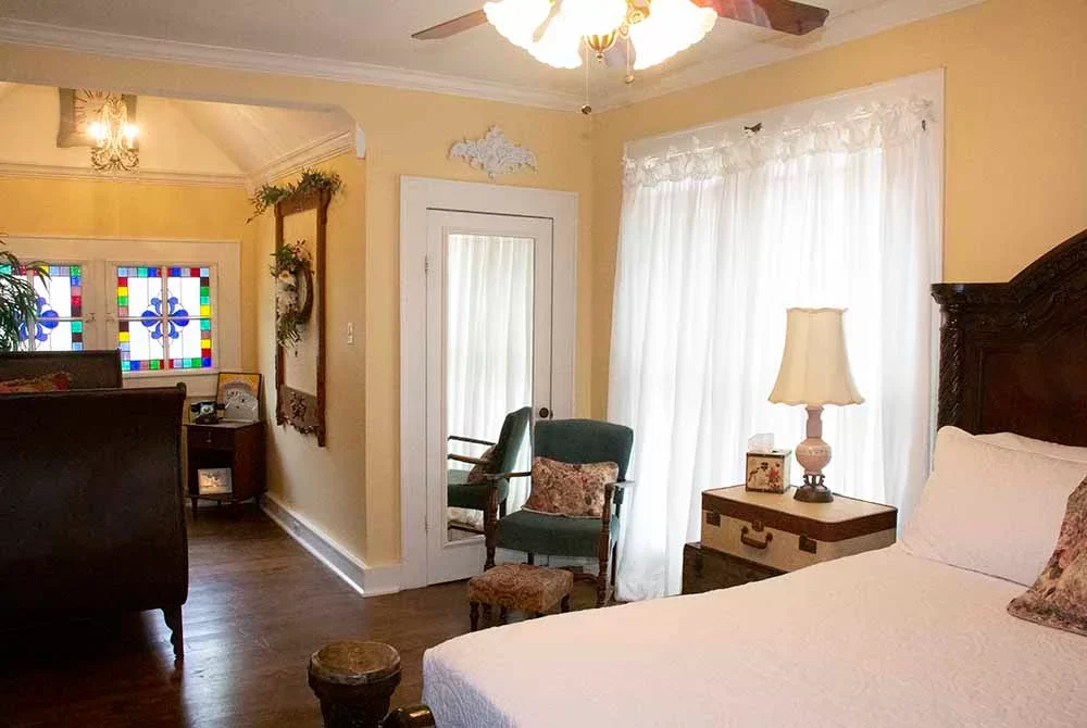 A sunny bedroom with soft yellow walls, a dark wood bed, sheer white curtains, and a seating area with stained glass windows and a private exit.