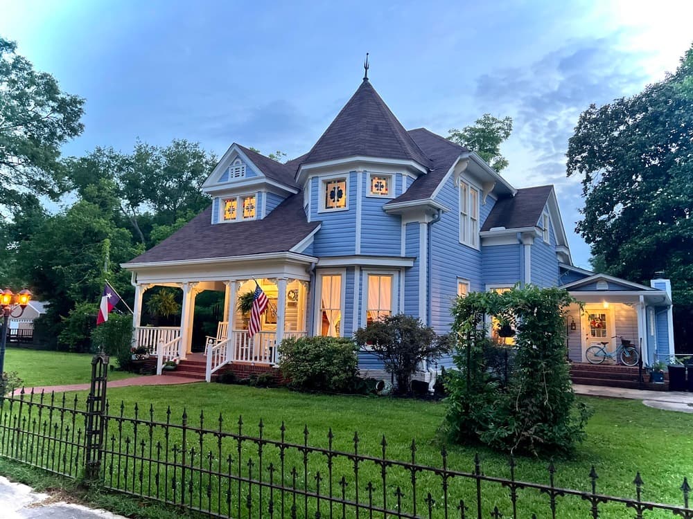 A charming blue Victorian house with a pointed roof, surrounded by greenery and a black iron fence.
