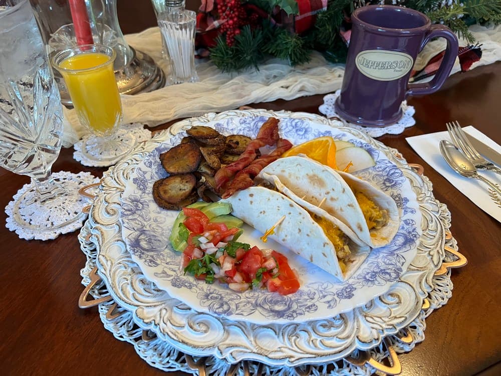 A beautifully arranged breakfast plate featuring tacos, bacon, fried plantains, fresh salsa, and sliced orange, alongside a glass of orange juice and a mug on a decorative table setting.
