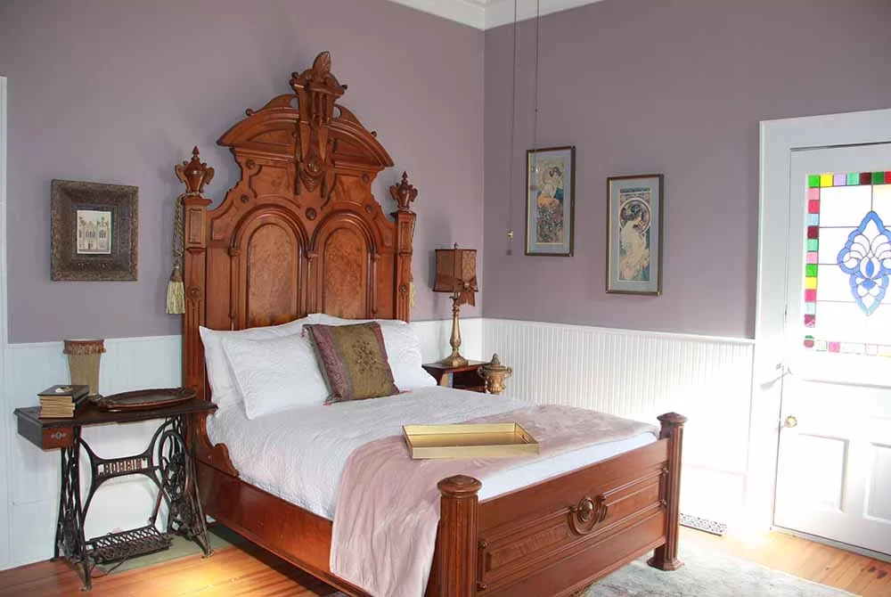 A bedroom featuring an antique wooden bed with a tall, ornate headboard, light purple walls, a vintage sewing machine table, and a door with stained glass.