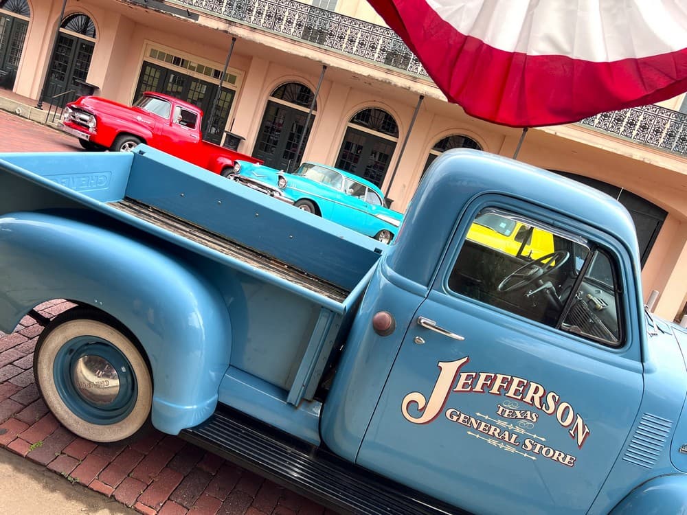 A vintage blue truck with "Jefferson, Texas General Store" featured prominently, parked in front of colorful classic cars.